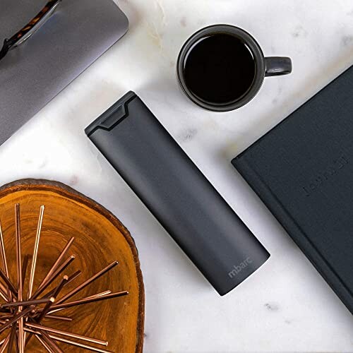 Black tumbler, coffee mug, and notebook on marble surface.