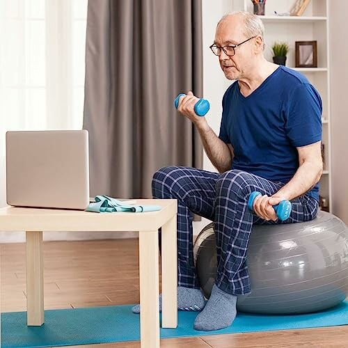 Senior man exercising with dumbbells on a stability ball in front of a laptop.